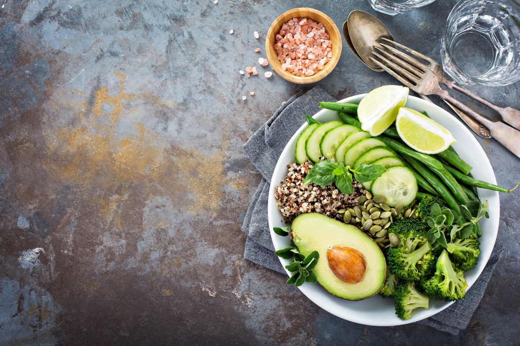 Overhead shot of a green vegan lunch bowl with quinoa, green beans, broccoli and avocado on a table with copy space