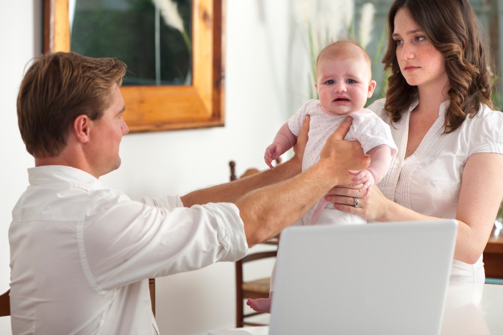 Frustrated father hands his crying baby to his wife, laptop in foreground